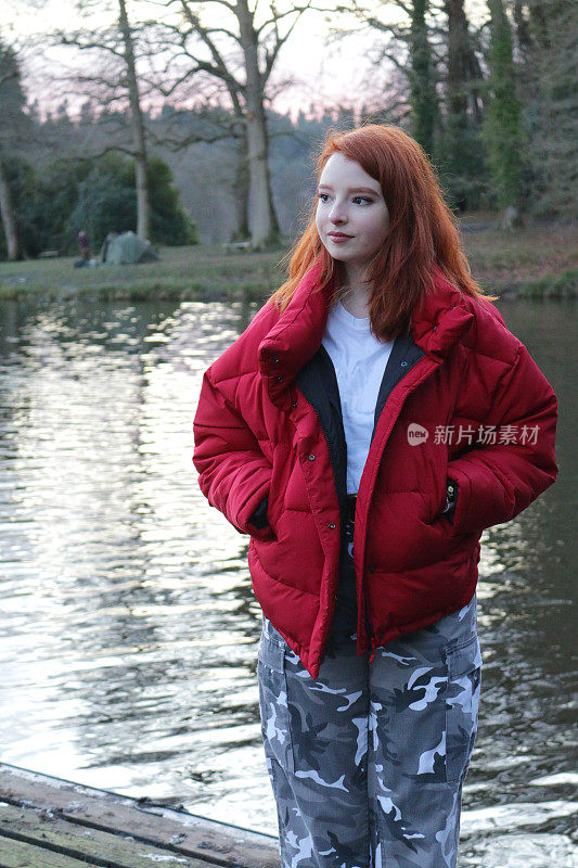 这是一个14 / 15岁的红发少女，皮肤苍白，脸上长着雀斑，穿着亮红色的羽绒服，外面罩着白色t恤和灰色迷彩长裤，站在户外的一个钓鱼湖旁边